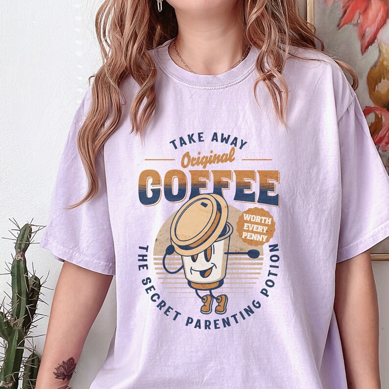 Op de afbeelding: Paars T-shirt met een retro-grafische afbeelding van een koffiebeker met de tekst "Take Away Original Coffee The Secret Parenting Potion Worth Every Penny".