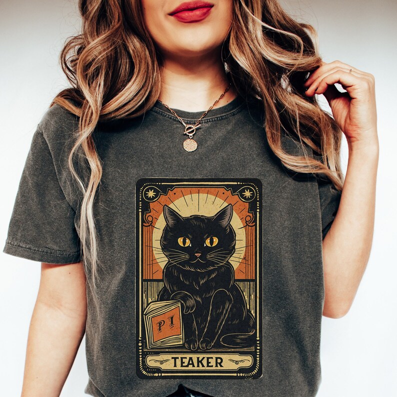 May include: A dark gray t-shirt with a vintage-style tarot card design featuring a black cat holding a book that says "PI" and the word "TEAKER" below the cat.