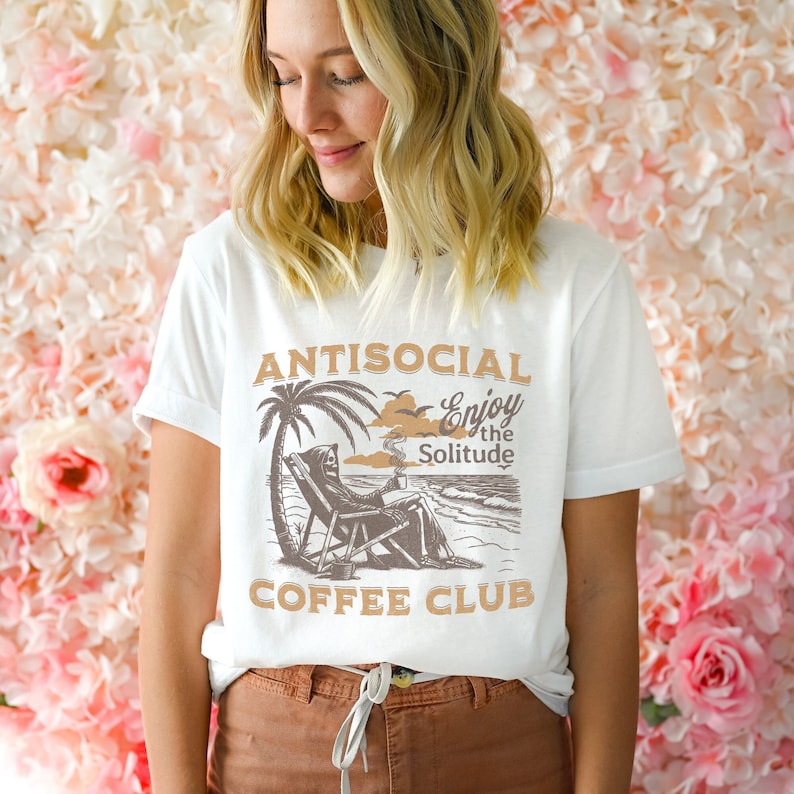 May include: White t-shirt featuring a graphic of a skeleton relaxing on a beach chair with a coffee cup. The text on the shirt reads "Antisocial Enjoy the Solitude Coffee Club".