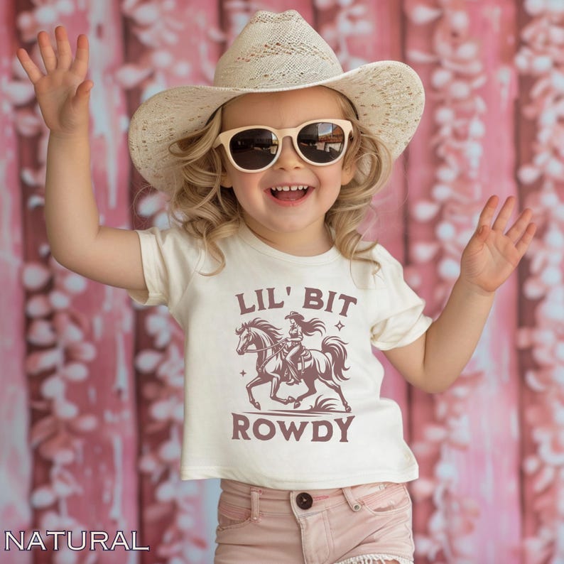 May include: A young child wearing a cream-colored t-shirt with the text "LIL' BIT ROWDY" and a graphic of a cowgirl on a horse. The child is also wearing a straw cowboy hat and sunglasses, with pink shorts. The background is a pink floral pattern.