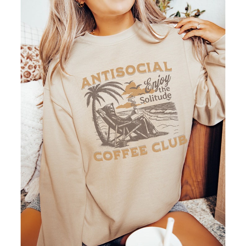 May include: A beige sweatshirt with a graphic print of a skeleton sitting on a beach chair with a palm tree and the text "ANTISOCIAL Enjoy the Solitude COFFEE CLUB".