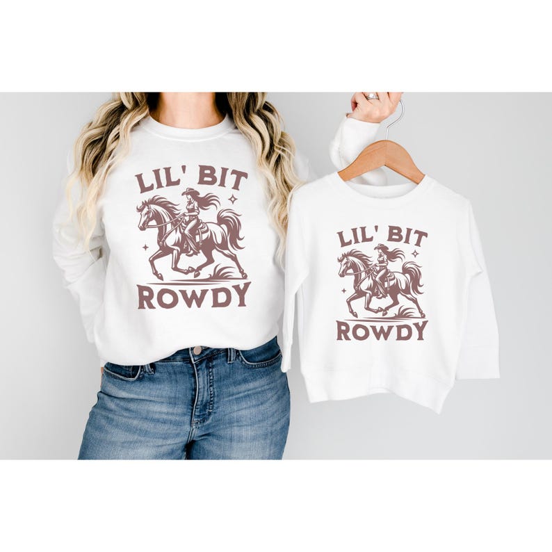 May include: White sweatshirts with a brown graphic of a cowgirl riding a horse, the text "LIL' BIT ROWDY" is above and below the graphic. One sweatshirt is worn, the other is on a wooden hanger.