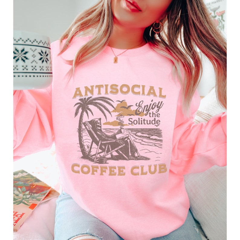 May include: Pink sweatshirt featuring a graphic of a skeleton relaxing in a beach chair with a cup of coffee. The text on the sweatshirt reads "Antisocial Enjoy the Solitude Coffee Club".