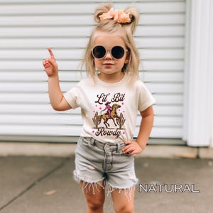 May include: A young girl wearing a light brown t-shirt with a graphic of a cowgirl riding a horse and the text "Lil' Bit Rowdy". She is wearing denim shorts and sunglasses. The background is a white brick wall.