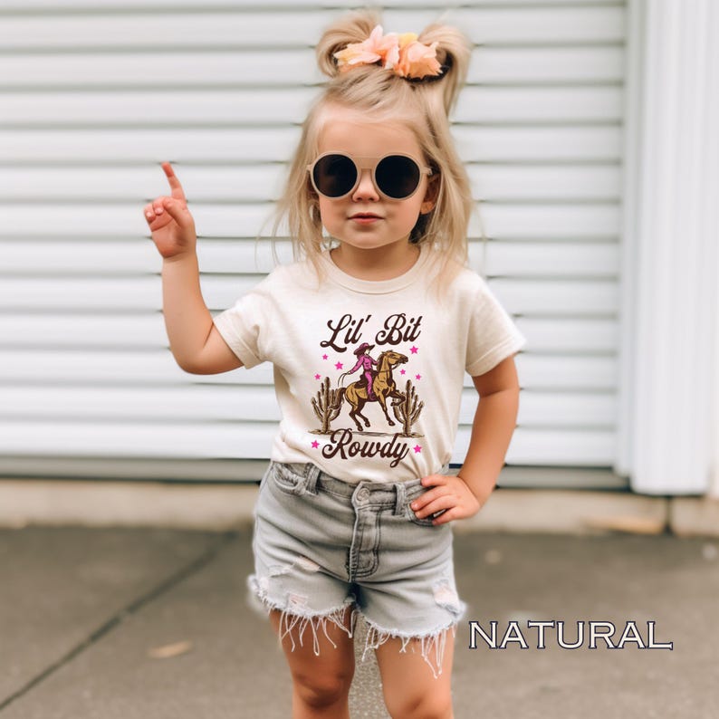 May include: A young girl wearing a light brown t-shirt with a graphic of a cowgirl riding a horse and the text "Lil' Bit Rowdy". She is wearing denim shorts and sunglasses. The background is a white brick wall.