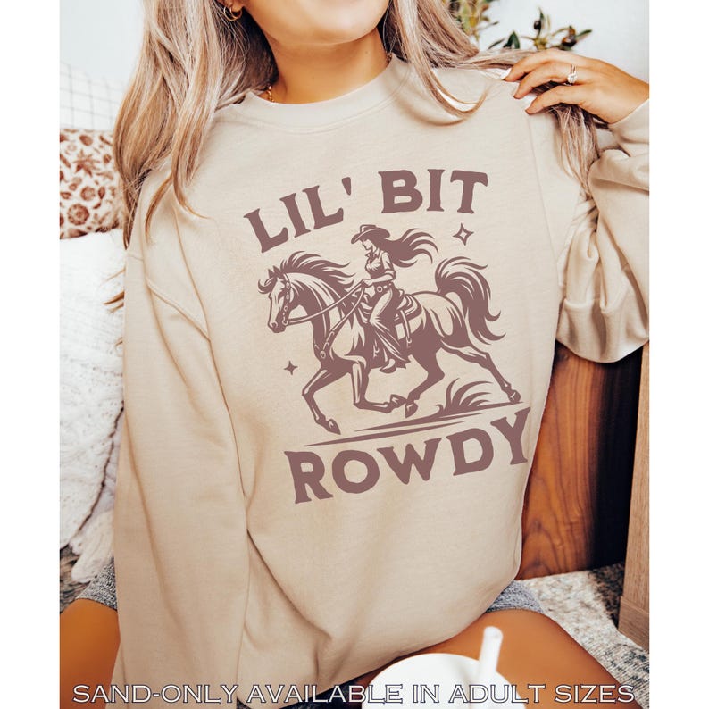 May include: A sand-colored sweatshirt with the text "LIL' BIT ROWDY" in brown. The graphic depicts a cowgirl riding a horse. The text "SAND-ONLY AVAILABLE IN ADULT SIZES" is at the bottom.