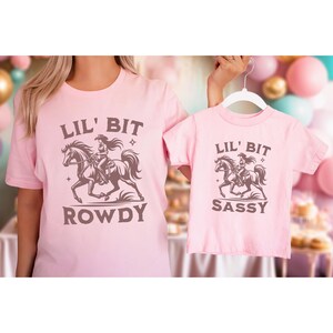 May include: Pink t-shirts with a brown graphic of a cowgirl riding a horse. The adult shirt says "Lil' Bit Rowdy" and the child's shirt says "Lil' Bit Sassy". The shirts are displayed on a light pink background.