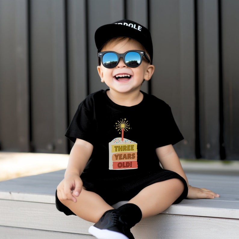 May include: A young child wearing a black t-shirt with a birthday cake graphic that says "Three Years Old!" The child is also wearing a black baseball cap with the word "DOLE" on it and black sunglasses.