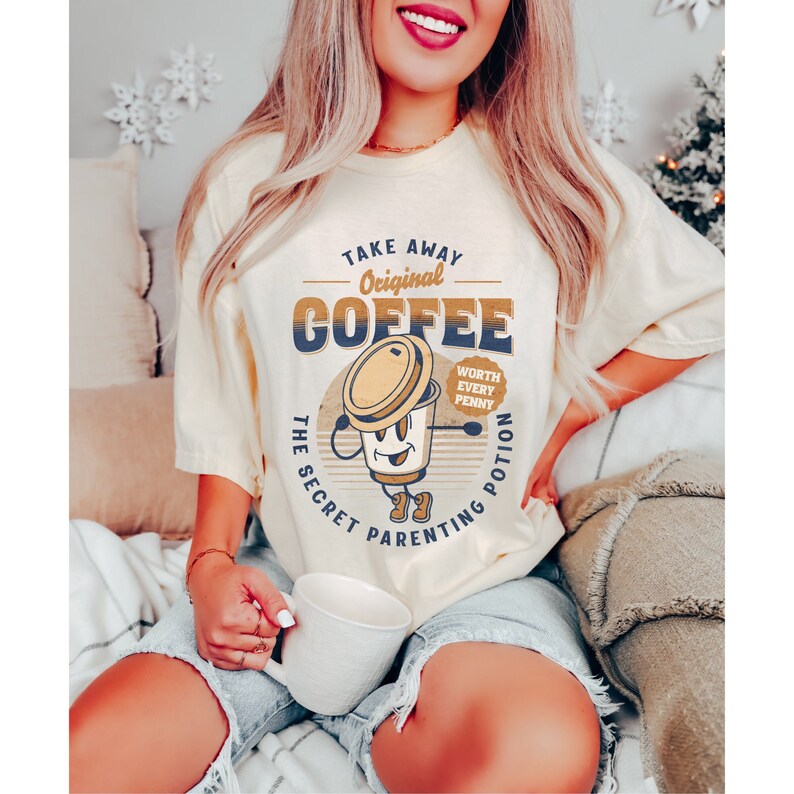 Op de afbeelding: Een lichtgeel T-shirt met een retro-grafische afbeelding van een koffiekopje met de tekst "Take Away Original Coffee The Secret Parenting Potion Worth Every Penny".