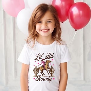 May include: A white t-shirt with a brown and pink graphic of a girl riding a horse with the text "Lil' Bit Rowdy" in brown letters.