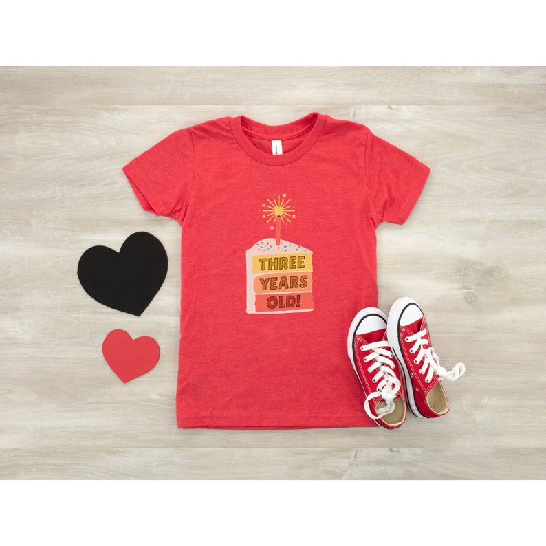 May include: Red t-shirt with a graphic of a layered cake with a lit candle and the text "THREE YEARS OLD!"