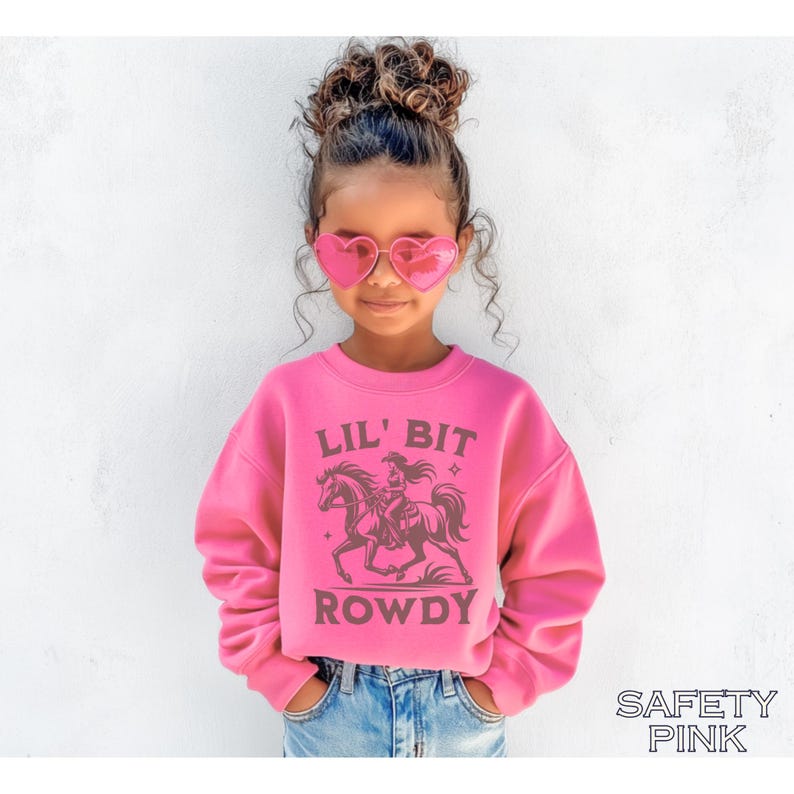 May include: A pink sweatshirt with the text "LIL' BIT ROWDY" and a graphic of a cowgirl riding a horse. The sweatshirt is a vibrant pink color and is paired with blue jeans. The words "SAFETY PINK" are printed at the bottom.
