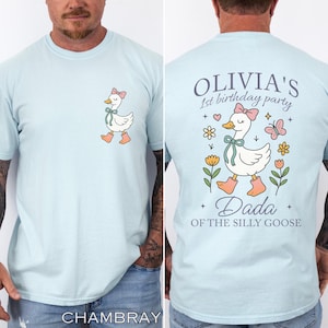 May include: Light blue t-shirt featuring a cartoon goose with a pink bow and boots. The back of the shirt has the text "Olivia's 1st birthday party" and "Dada of the silly goose" with floral and butterfly graphics.