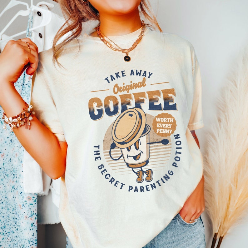 Op de afbeelding: Een lichtbeige t-shirt met een retro-stijl grafische afbeelding van een koffiebeker met de tekst "Take Away Original Coffee The Secret Parenting Potion Worth Every Penny".