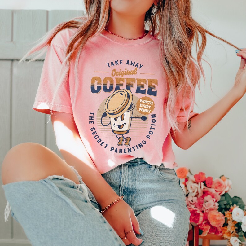 Op de afbeelding: Een roze T-shirt met een afbeelding van een koffiebeker met een gezicht en de tekst "Take Away Original Coffee The Secret Parenting Potion Worth Every Penny".