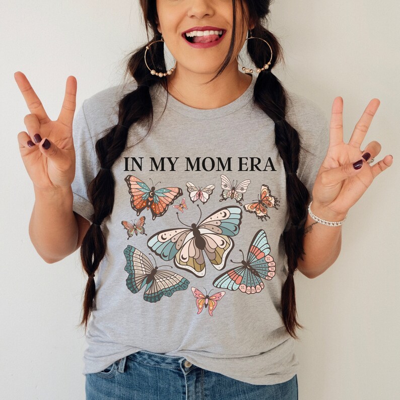 K&ouml;nnte beinhalten: Ein graues T-Shirt mit dem Text "In My Mom Era" und einem bunten Schmetterlingsdesign. Die Schmetterlinge sind in Blaut&ouml;nen, Gr&uuml;n, Rosa und Orange.