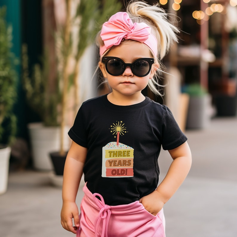 May include: A black t-shirt with a colorful birthday cake graphic and the text "THREE YEARS OLD!" on it. The shirt is being worn by a young child wearing pink sunglasses and a pink headband.