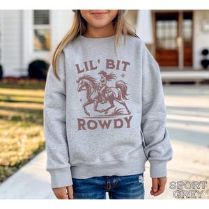 May include: A light gray sweatshirt with the words "LIL' BIT ROWDY" in brown lettering. The sweatshirt features a graphic of a cowgirl riding a horse. The words "SPORT GREY" are printed at the bottom right.