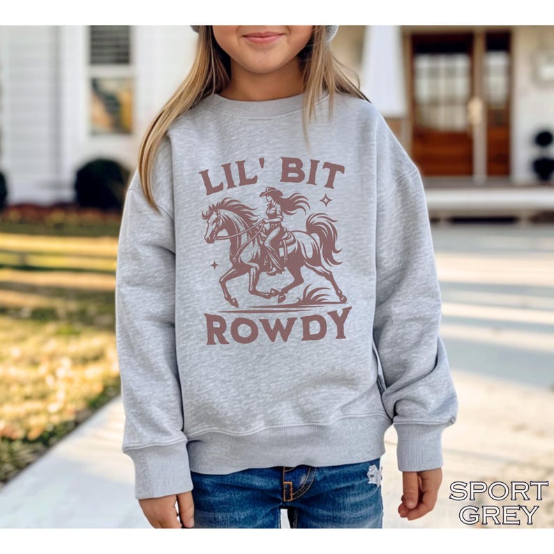 May include: A light gray sweatshirt with the words "LIL' BIT ROWDY" in brown lettering. The sweatshirt features a graphic of a cowgirl riding a horse. The words "SPORT GREY" are printed at the bottom right.