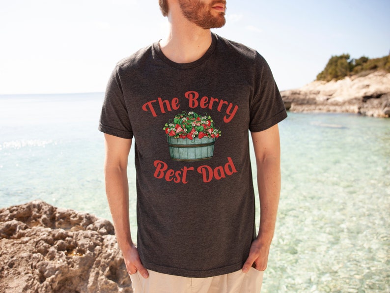May include: A dark grey t-shirt with the text "The Berry Best Dad" in red lettering. A cartoon illustration of a bucket overflowing with red strawberries is above the text.