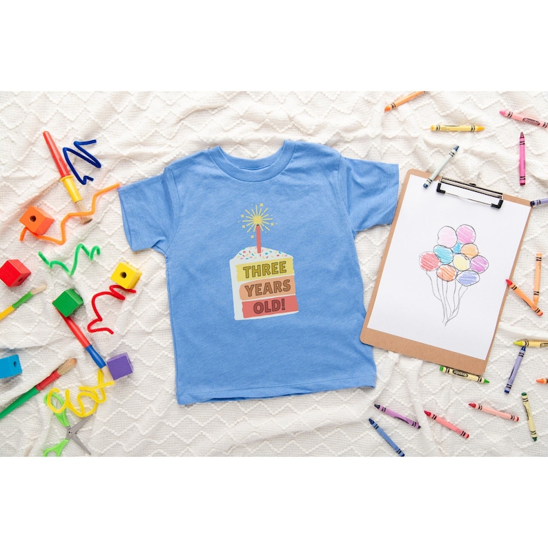 May include: A light blue t-shirt with a colorful birthday cake graphic that says "Three Years Old!" on it. The shirt is surrounded by crayons and other children's toys.