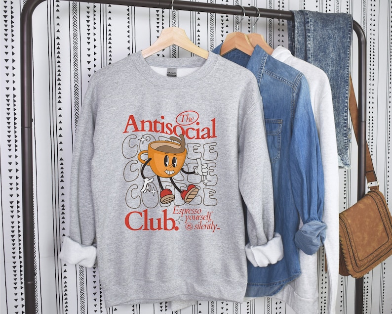 K&ouml;nnte beinhalten: Grauer Pullover mit einem Comic-Kaffeebecher-Motiv und dem Text "The Antisocial Club. Espresso yourself, silently."