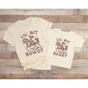 May include: Two cream-colored t-shirts with short sleeves. The larger shirt has the text "LIL' BIT ROWDY" with a graphic of a cowgirl riding a horse. The smaller shirt says "LIL' BIT SASSY" with a similar graphic.