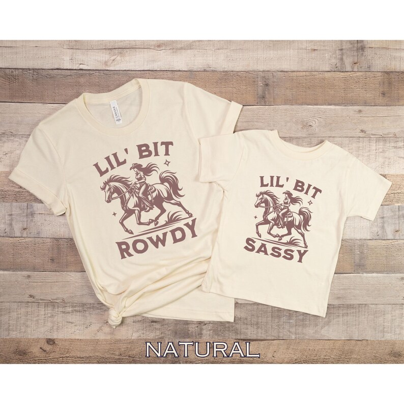 May include: Two cream-colored t-shirts with short sleeves. The larger shirt has the text "LIL' BIT ROWDY" with a graphic of a cowgirl riding a horse. The smaller shirt says "LIL' BIT SASSY" with a similar graphic.