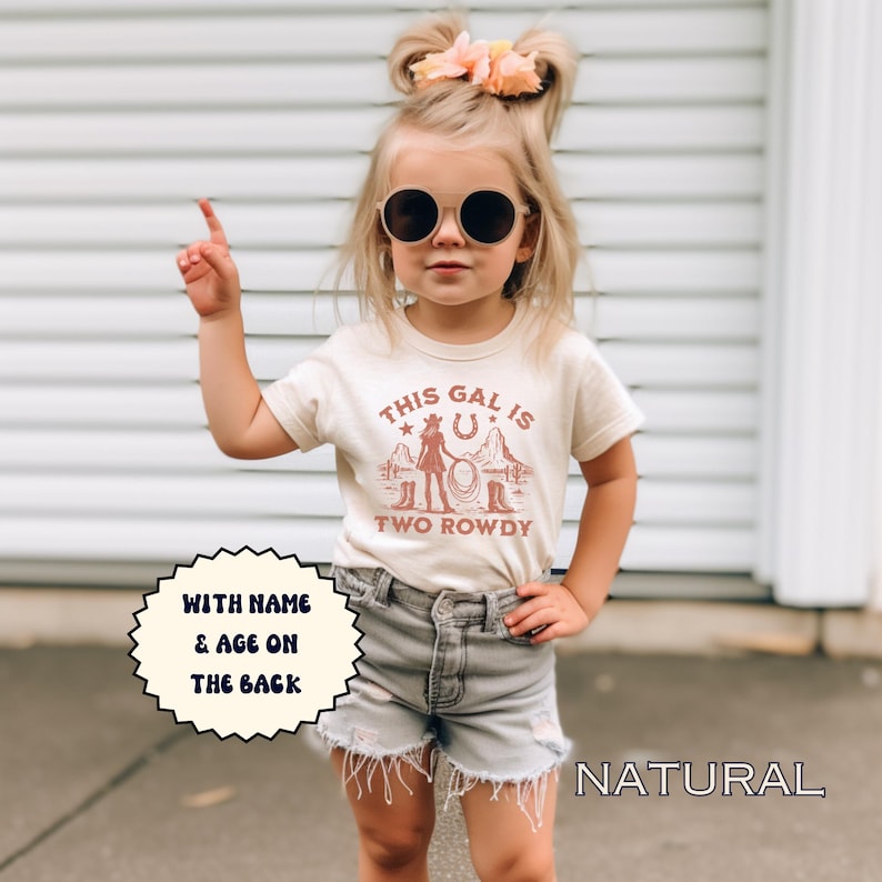 Puede incluir: Una camiseta beige claro con un gr&aacute;fico marr&oacute;n de una vaquera con un lazo y el texto "This gal is two rowdy". La camiseta tiene mangas cortas y cuello redondo. El texto "With name & age on the back" est&aacute; en un c&iacute;rculo blanco en el lado izquierdo de la imagen. El texto "NATURAL" est&aacute; en el lado derecho de la imagen.