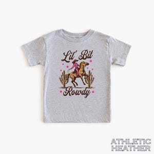 May include: A gray t-shirt with a graphic of a cowgirl riding a horse with the text "Lil' Bit Rowdy" in brown lettering and pink stars.