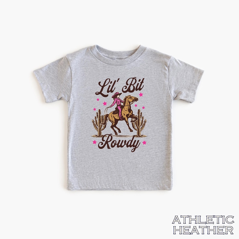 May include: A gray t-shirt with a graphic of a cowgirl riding a horse with the text "Lil' Bit Rowdy" in brown lettering and pink stars.