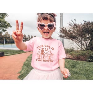 Puede incluir: Una camiseta rosa con un gr&aacute;fico marr&oacute;n de una ni&ntilde;a con un sombrero de vaquero y botas. El texto en la camiseta dice "This gal is two rowdy".