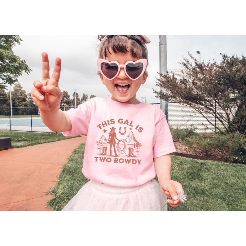 Puede incluir: Una camiseta rosa con un gr&aacute;fico marr&oacute;n de una ni&ntilde;a con un sombrero de vaquero y botas. El texto en la camiseta dice "This gal is two rowdy".