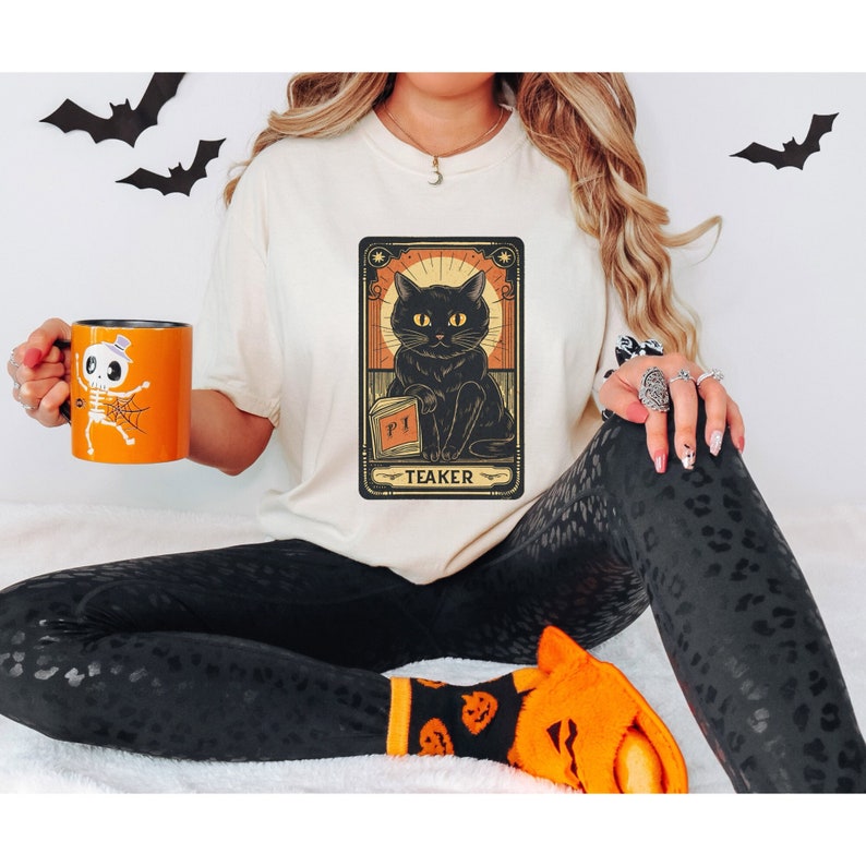 May include: A white t-shirt with a black cat illustration in a vintage tarot card style. The card reads "Teaker" and features a black cat holding a book with the letter "PI" on the cover.