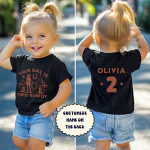 Puede incluir: Camiseta negra con un gr&aacute;fico marr&oacute;n de una ni&ntilde;a con un lazo y el texto "This gal is two rowdy". La parte posterior de la camiseta tiene una estrella marr&oacute;n, el nombre "Olivia" y el n&uacute;mero "2" en marr&oacute;n.