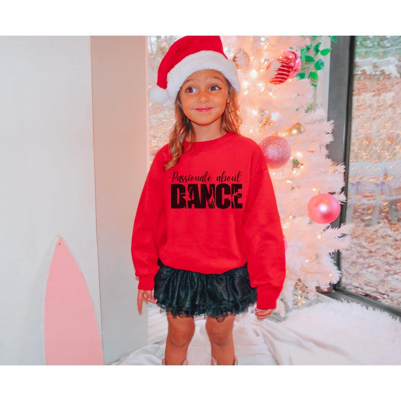 May include: A young person wearing a red sweatshirt with the text "Passionate about DANCE" in black. They are wearing a black tulle skirt and a red Santa hat. The person is standing in front of a white Christmas tree.