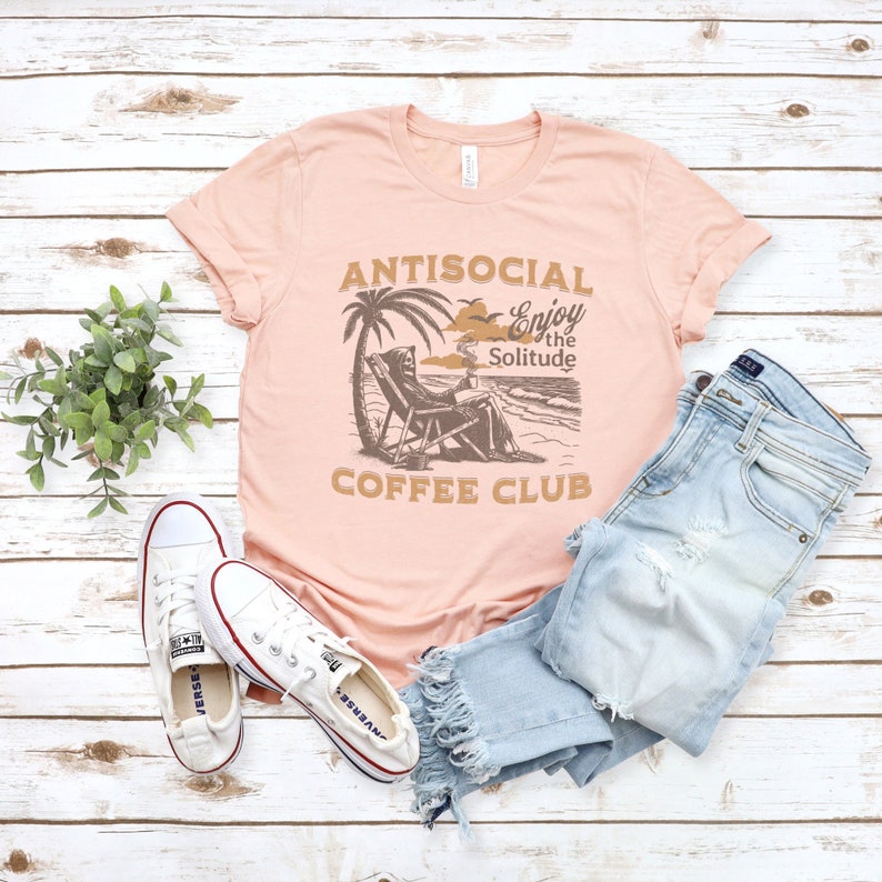 May include: A pink t-shirt featuring the text "ANTISOCIAL Enjoy the Solitude COFFEE CLUB" and a graphic of a skeleton sitting on a beach chair with a cup of coffee. The skeleton is wearing a hooded robe and has a skull face. The graphic is in brown and black.