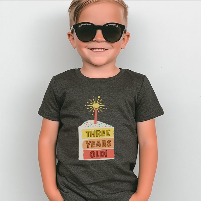 May include: A dark gray t-shirt with a colorful birthday cake graphic. The cake has a lit candle and the text "THREE YEARS OLD!" on it.