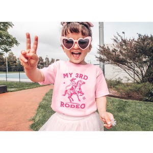 May include: A pink t-shirt with a pink graphic of a girl riding a horse. The graphic says "My 3rd Rodeo".