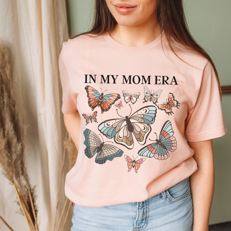 K&ouml;nnte beinhalten: Ein rosa T-Shirt mit dem Text "In My Mom Era" und einem Design aus bunten Schmetterlingen.