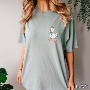 May include: A light green t-shirt featuring a cartoon goose with a pink bow, blue ribbon, and pink boots. The goose is walking. The shirt is a solid color and has short sleeves.