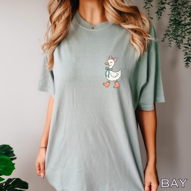 May include: A light green t-shirt featuring a cartoon goose with a pink bow, blue ribbon, and pink boots. The goose is walking. The shirt is a solid color and has short sleeves.