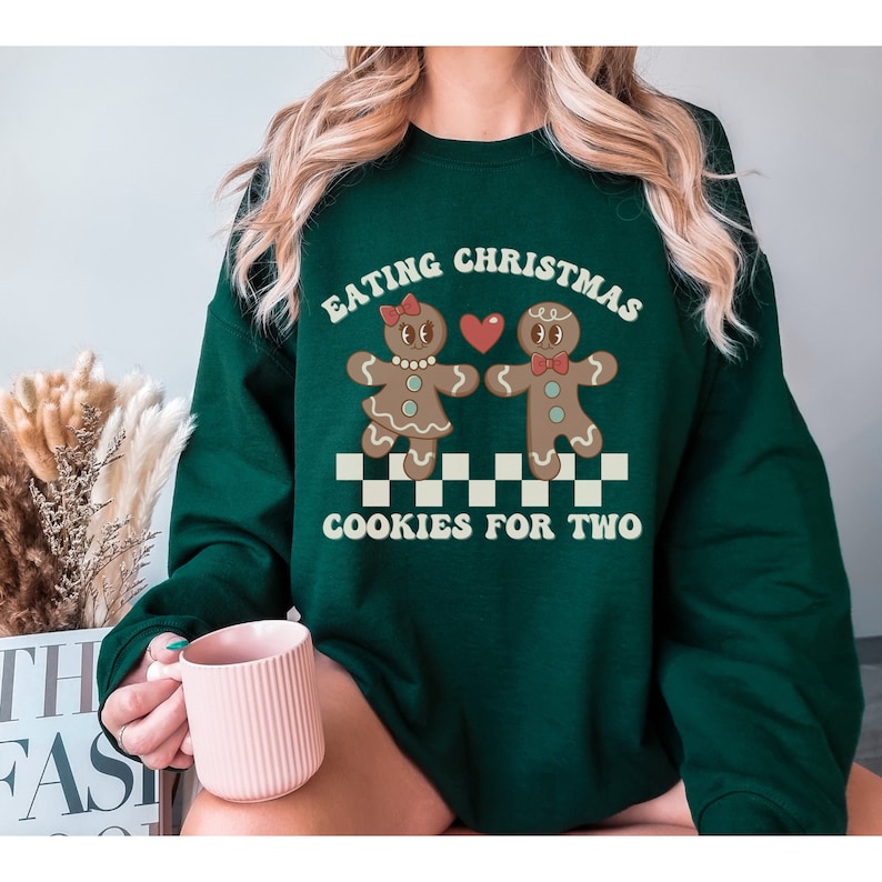 May include: Green Christmas sweater with a graphic of two gingerbread people holding hands and a heart. The text on the sweater reads "Eating Christmas Cookies for Two."