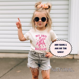 May include: A young girl wearing a white t-shirt with pink text that says "My 3rd Rodeo" and a pink graphic of a cowboy riding a horse. The girl is wearing denim shorts and sunglasses. The text "Add name & age on the back!" is in a white circle with a pink outline. The text "NATURAL" is in the bottom right corner of the image.