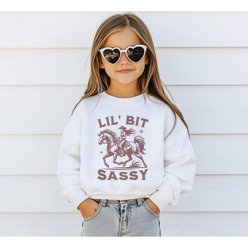May include: A white sweatshirt with the text "LIL' BIT SASSY" and a graphic of a cowgirl riding a horse. The girl in the image is wearing heart-shaped sunglasses and blue jeans.