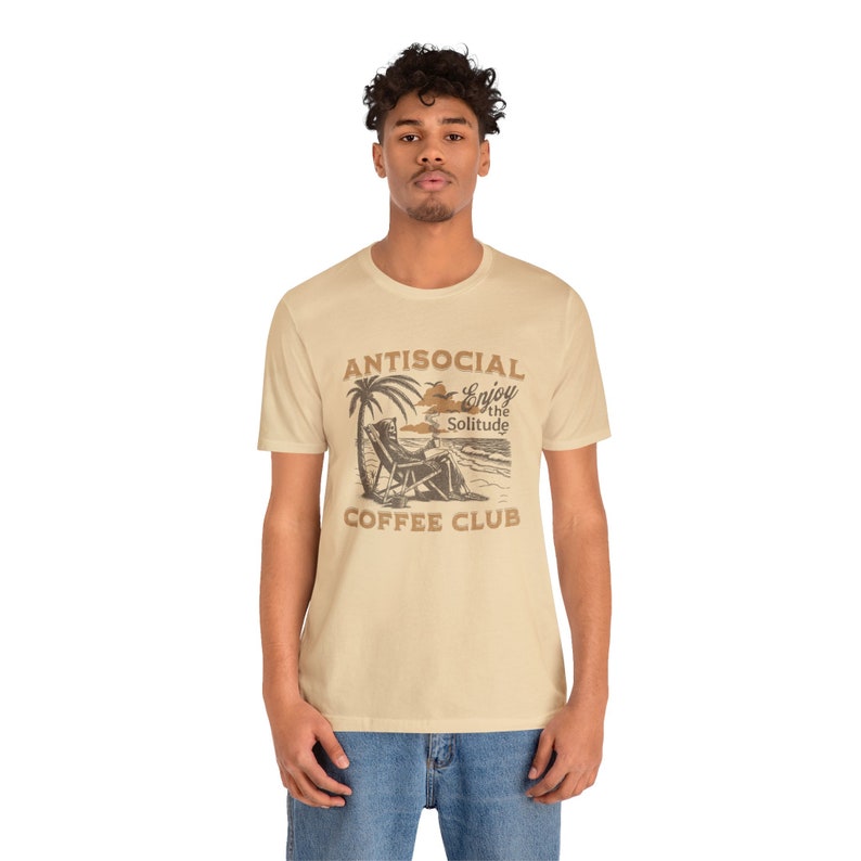 May include: A beige t-shirt featuring a brown graphic of a person relaxing in a beach chair beneath a palm tree. The graphic reads "Antisocial Enjoy the Solitude Coffee Club".