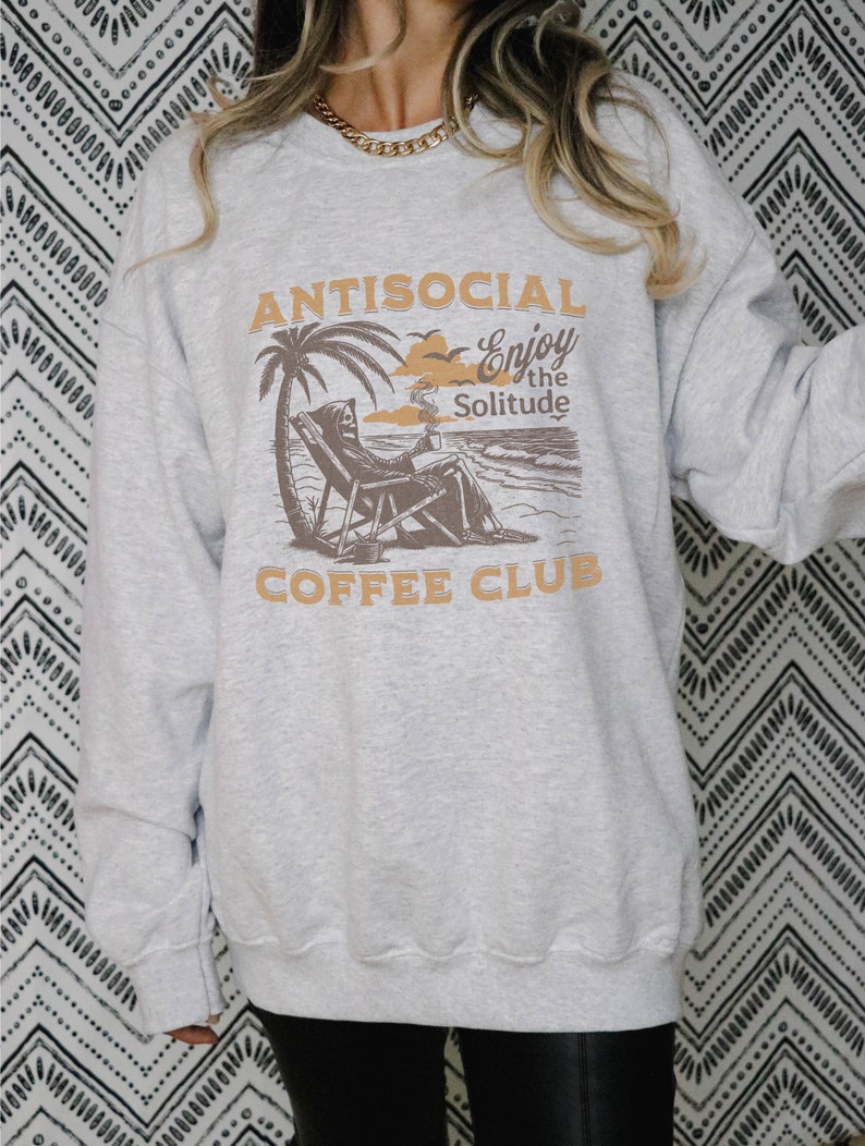 May include: Grey sweatshirt featuring a graphic of a skeleton sitting on a beach chair with a cup of coffee. The text on the sweatshirt reads "Antisocial Enjoy the Solitude Coffee Club".