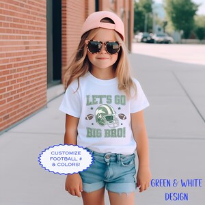 Puede incluir: Una camiseta blanca con el texto "LET'S GO BIG BRO!" en verde, con un casco de f&uacute;tbol americano y balones de f&uacute;tbol. La camiseta tambi&eacute;n tiene el texto "CUSTOMIZE FOOTBALL # & COLORS!" y "GREEN & WHITE DESIGN". El ni&ntilde;o lleva pantalones cortos vaqueros azules y una gorra rosa.