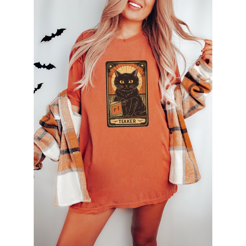 May include: A burnt orange t-shirt with a black cat illustration. The cat is holding a book with the word "PI" on it. The illustration is surrounded by a gold frame with the word "TEAKER" at the bottom.