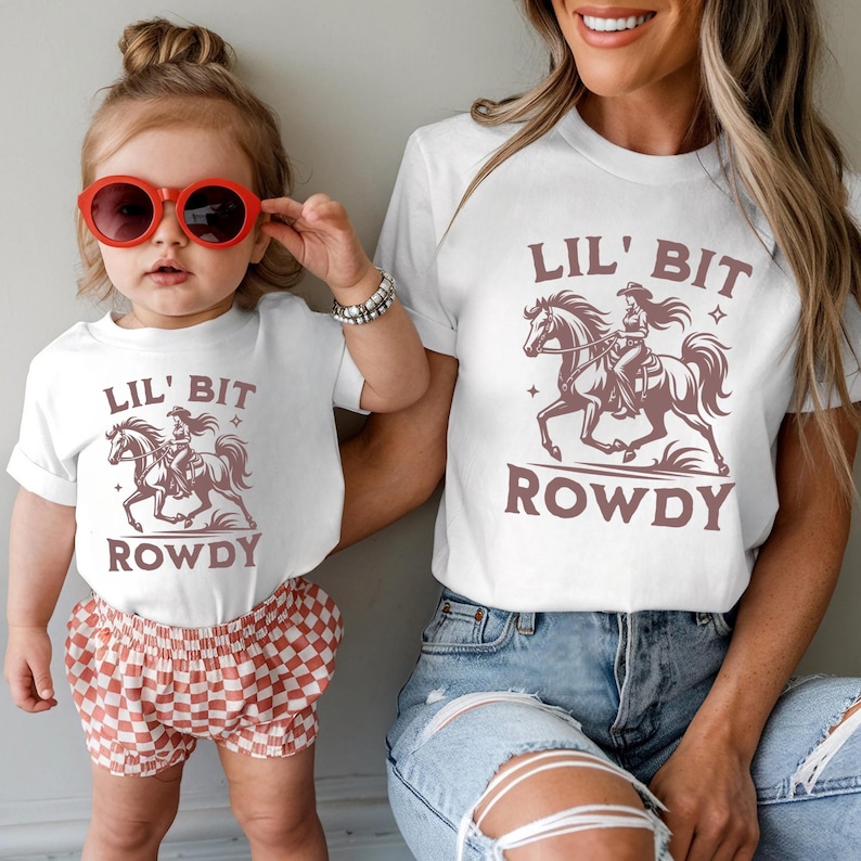 May include: White t-shirts with a brown graphic of a cowgirl riding a horse, the text "Lil' Bit Rowdy" is above and below the graphic. The child is wearing red sunglasses and checkered shorts. The adult is wearing blue jeans.